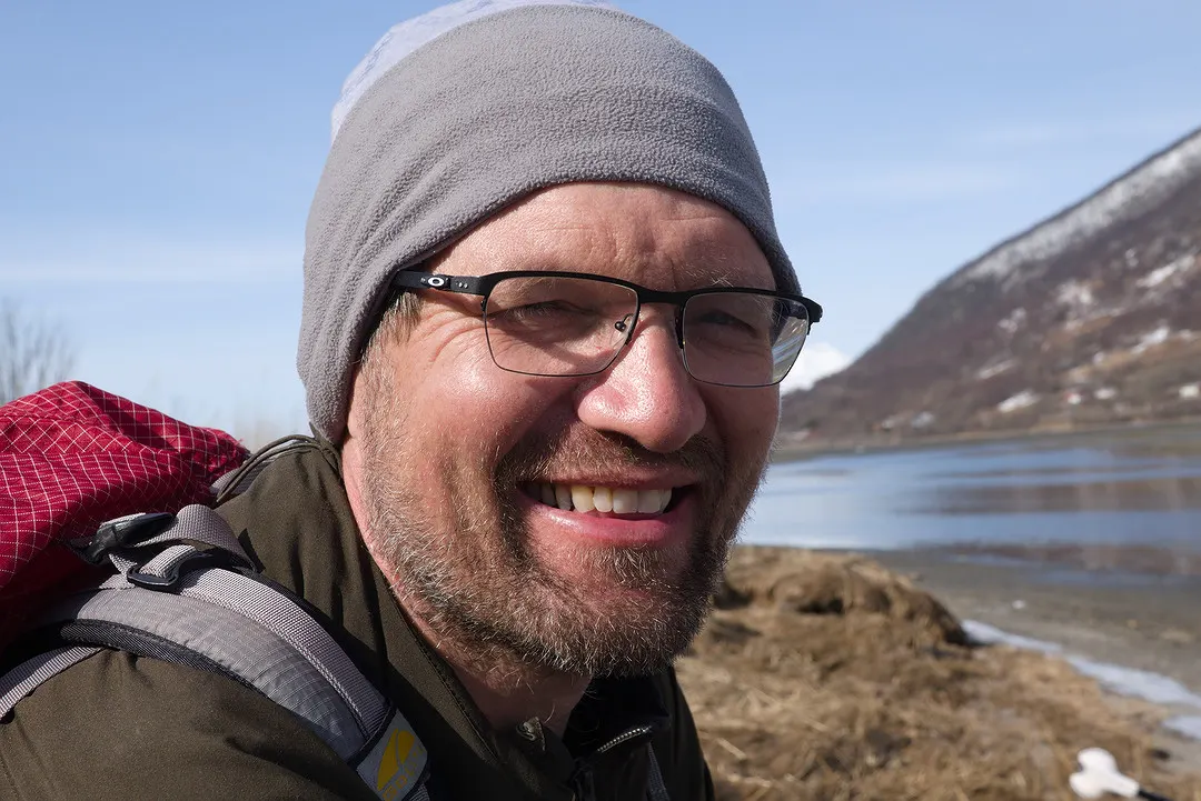 Roy smiling in a beanie by a fjord on a sunny spring day